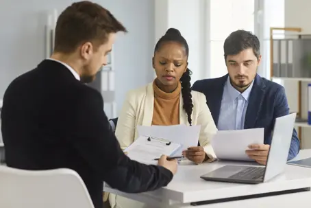 Serious married couple carefully reads paper documents sitting with a bank agent in the office. Caucasian husband and his dark skinned wife check bills, contract terms, mortgage or loan agreemen