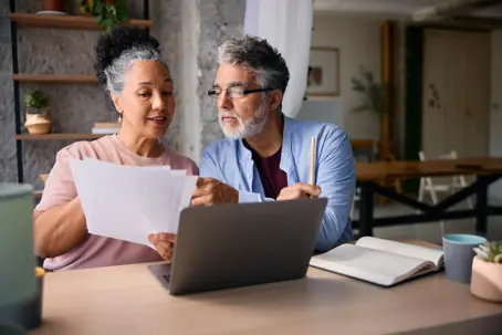 Senior couple sitting together at a dining table, reviewing paperwork and using a laptop, engaged in serious discussions about finances and retirement planning in their cozy home